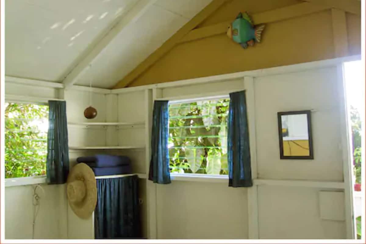A room with a sloped ceiling, a window with blue curtains, a wall-mounted mirror, and a hat hanging on the wall.