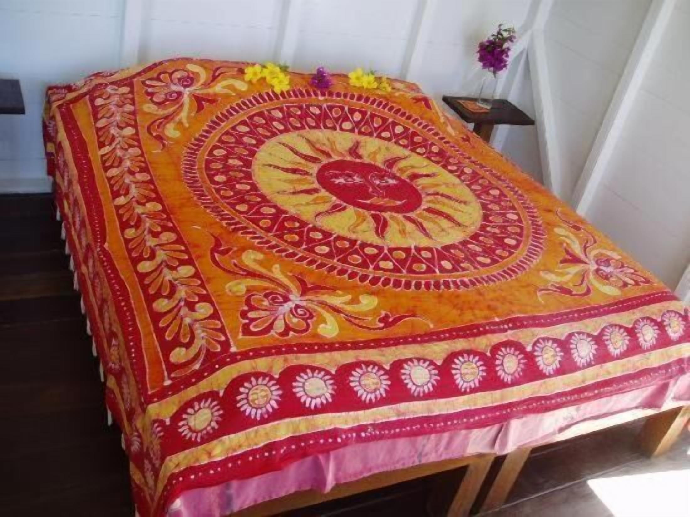 A bed with a red and yellow patterned bedspread, a wooden headboard, and a small table with a vase of flowers.