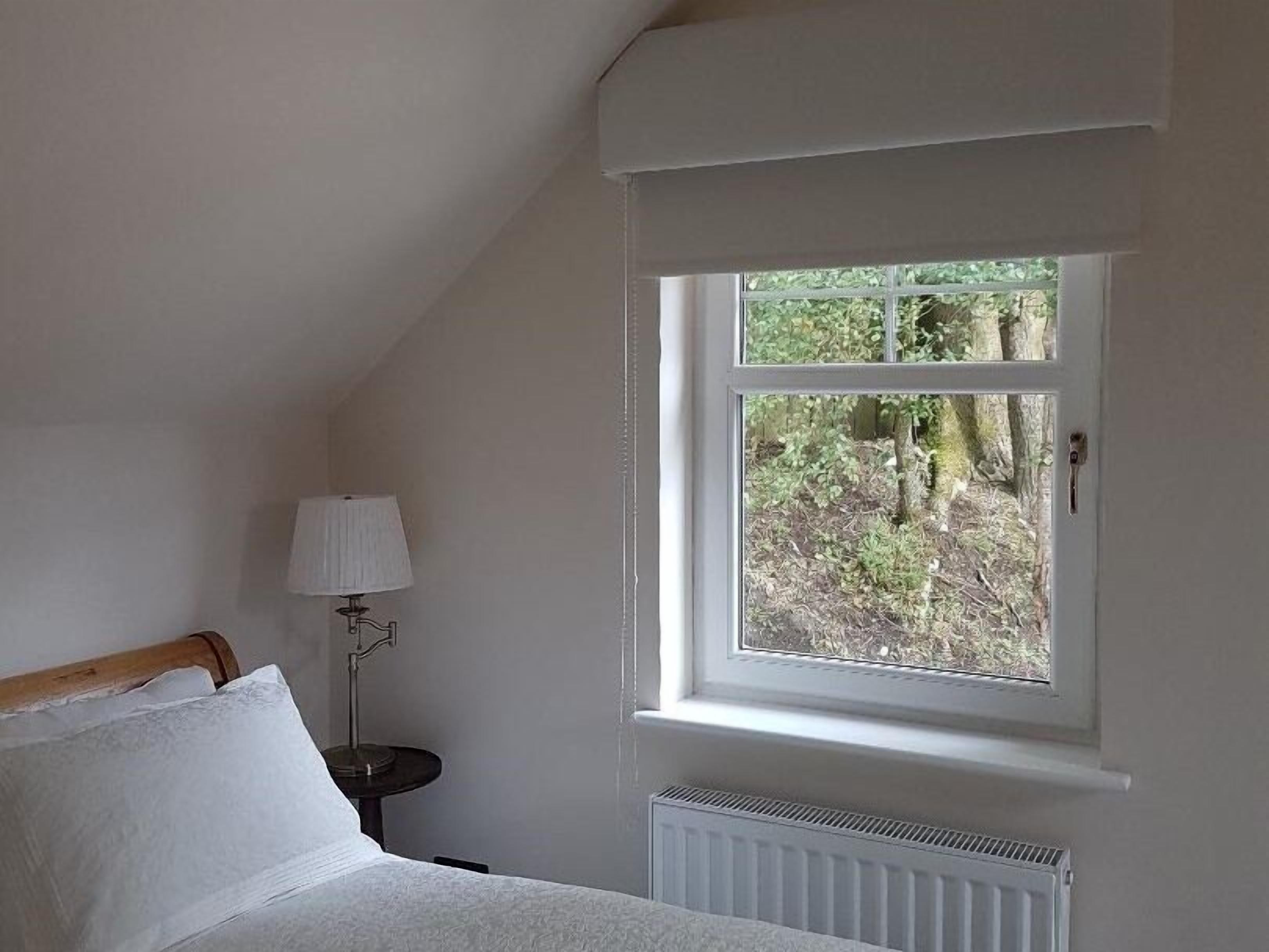 Un dormitorio con cama, mesita de noche, lámpara y ventana con vistas a árboles.