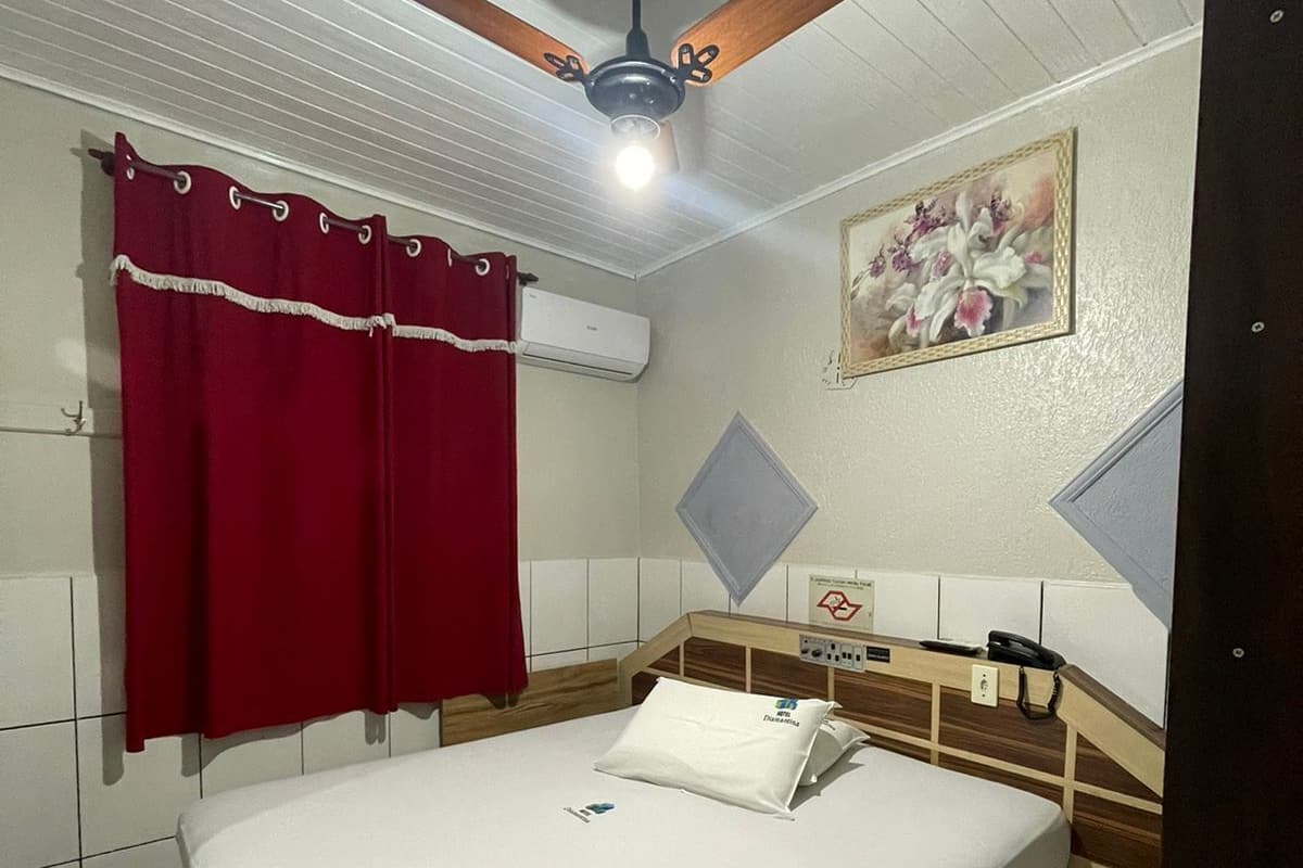 A hotel room with a bed, a ceiling fan, a red curtain, an air conditioning unit, a bedside table with a phone, and a painting on the wall.