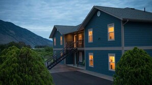 A two-story building with a staircase leading to the upper floor, surrounded by greenery and mountains in the background. - Klippers Guest Suites (Keremeos)