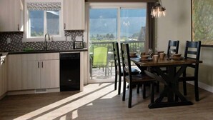 A modern kitchen with a dishwasher, a dining area with a table set for four, and a view of the outdoors through a sliding door. - Klippers Guest Suites (Keremeos)