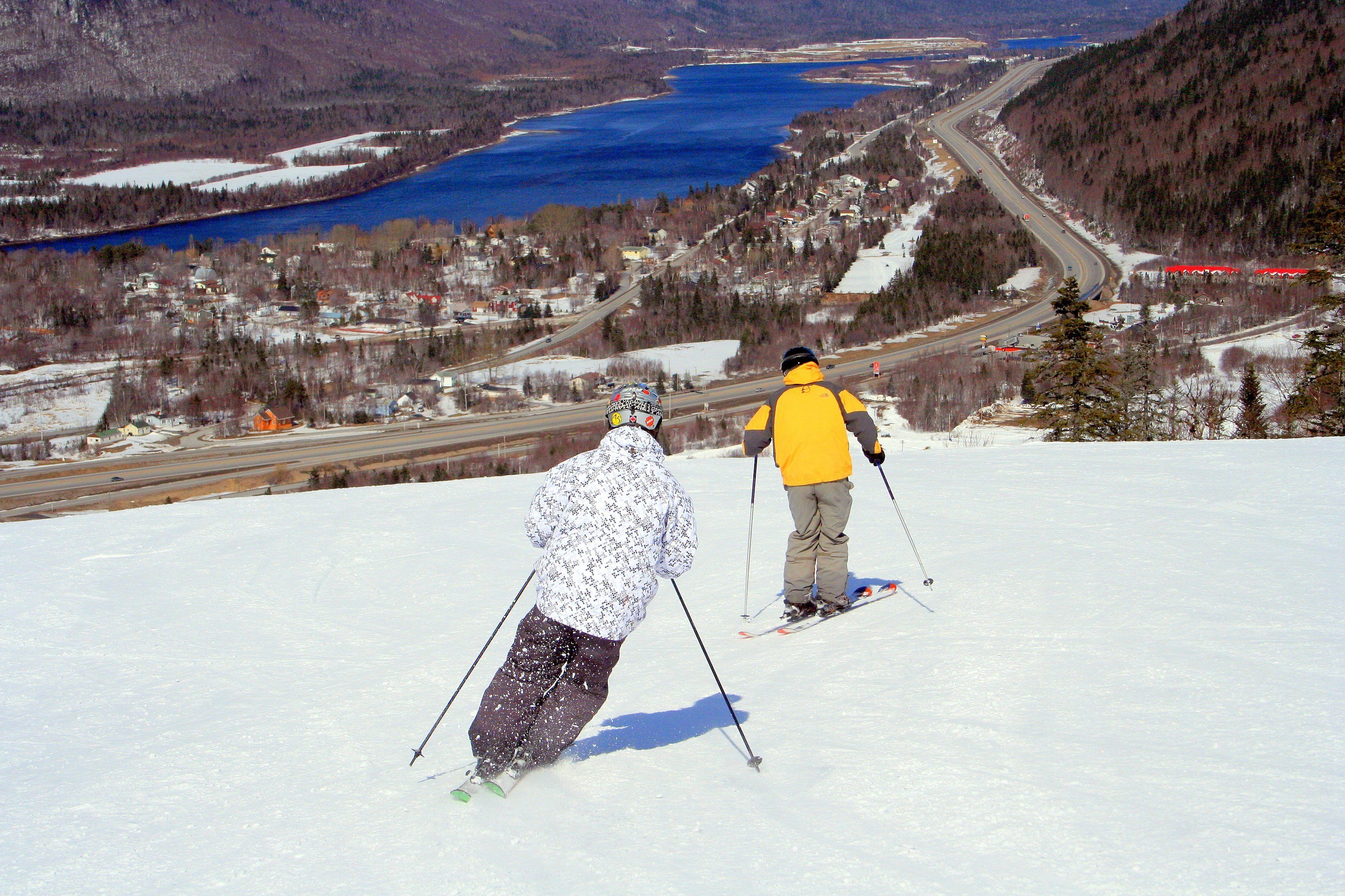 Skiers on a snowy slope with a view of a lake and a highway in the distance.