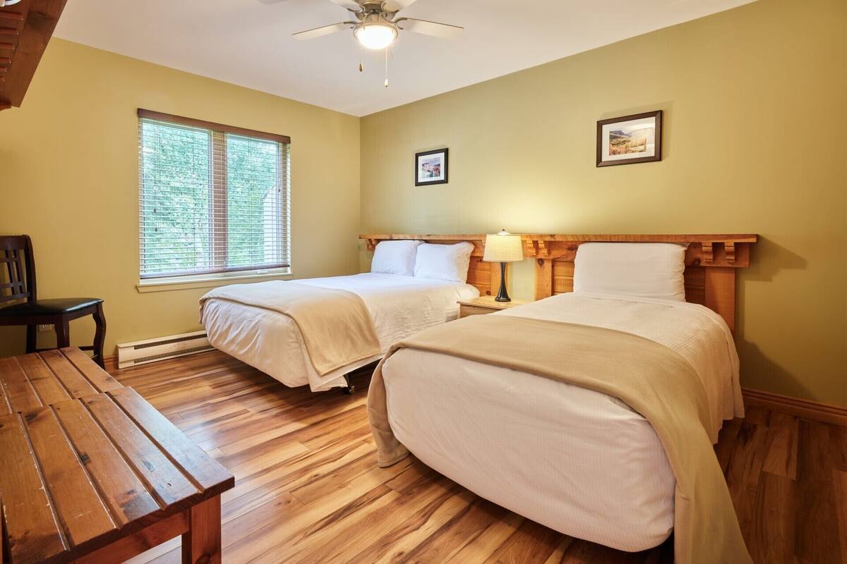 Een kamer met twee bedden, een houten bankje, een plafondventilator en een raam met jaloezieën.
