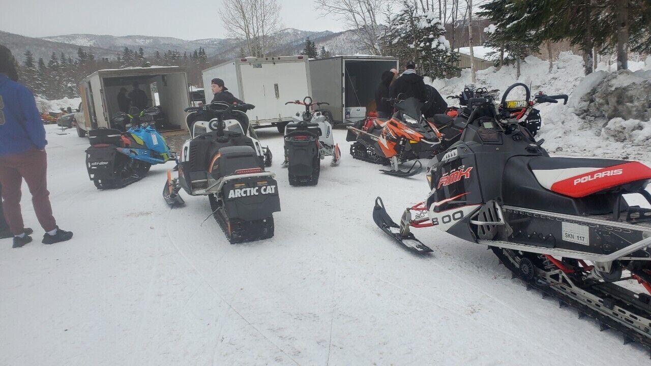 A group of snowmobiles parked in a snowy area with a trailer and a person standing nearby.