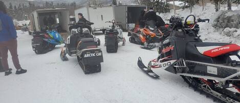 Un grupo de motos de nieve estacionadas en una zona nevada, con un remolque y una persona de pie cerca.