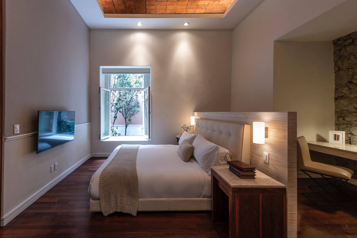 Une chambre à coucher comprenant un lit, une table de chevet, une lampe, une télévision et une fenêtre donnant sur un arbre.
