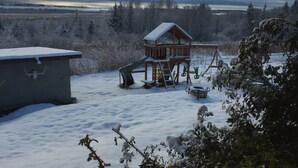 A wooden cabin with a snow-covered roof, a slide, and a deck, surrounded by snow-covered trees and a distant view of a lake. - Homer Stay and Play (Homer)
