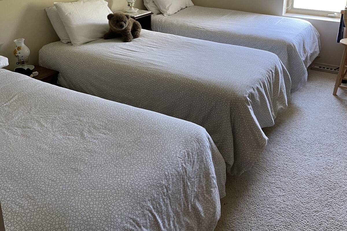 A room with three beds, each with a white bedspread and pillows. There is a small table with a lamp and a teddy bear on one bed. The room has a sloped ceiling and a window with blinds.