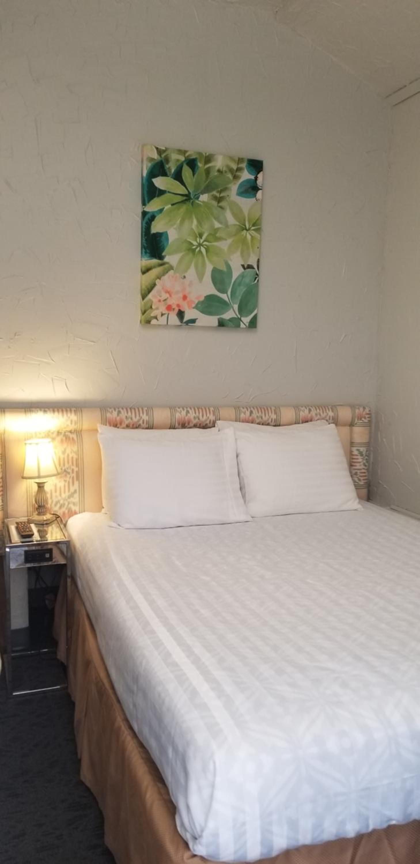 Un lit bien fait, recouvert d’une couvre-lit blanche et agrémenté de deux oreillers, une lampe de chevet et un tableau représentant des feuilles tropicales, accroché au mur.