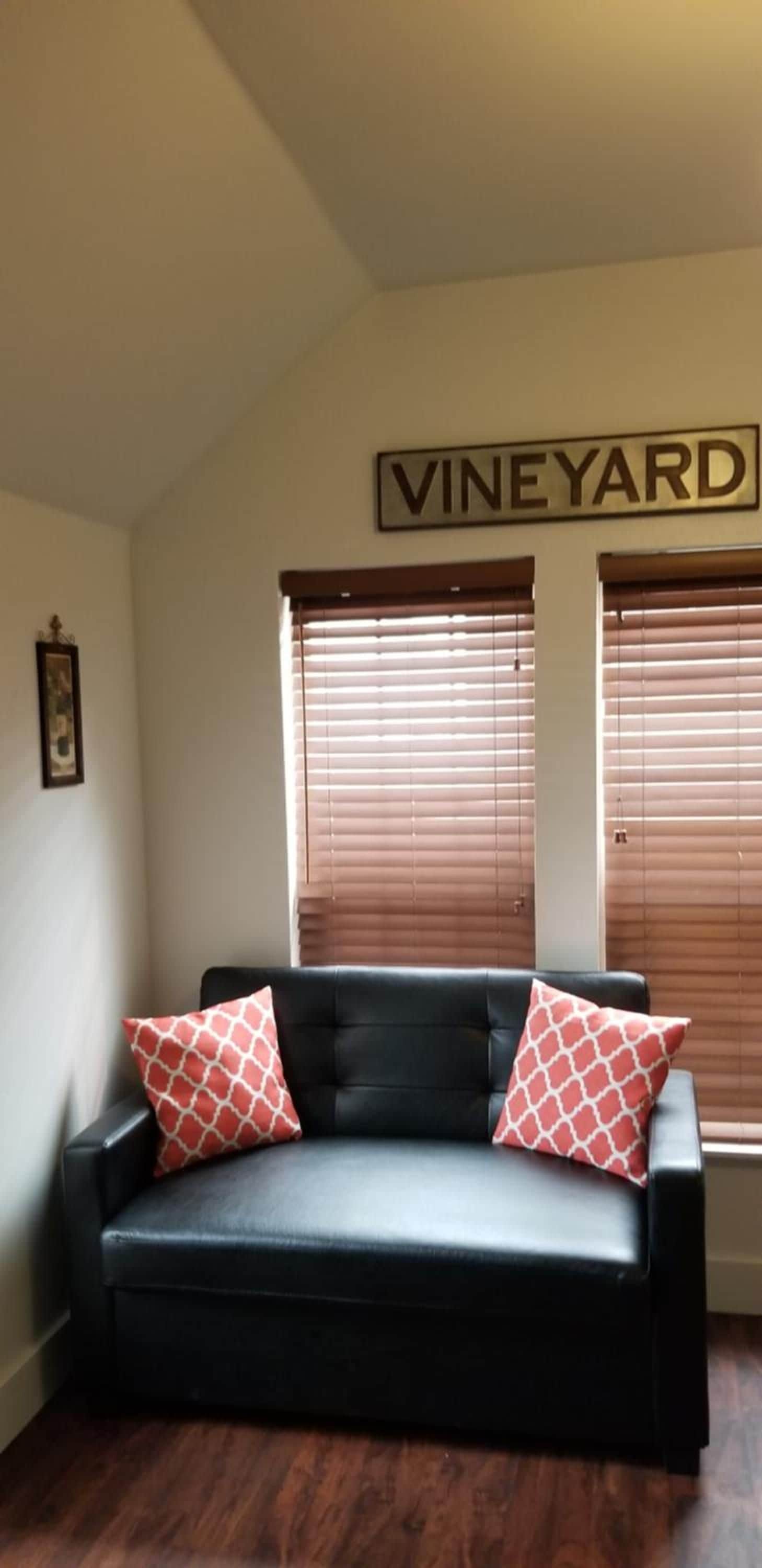 A black leather sofa with two patterned pillows, a framed picture on the wall, and a sign that reads 'VINEYARD' above two windows with blinds.