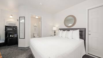 A hotel room with a large bed, a bedside lamp, a mirror, a door, and a kitchenette with a microwave.