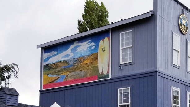 A building with a mural of a landscape and an apple, outdoor seating with red chairs and pink umbrellas, and a clock on the building. - Deep Water Inn (Chelan)