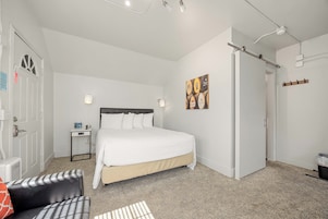 Un dormitorio con cama, mesita de noche, lámpara y una obra de arte en la pared.