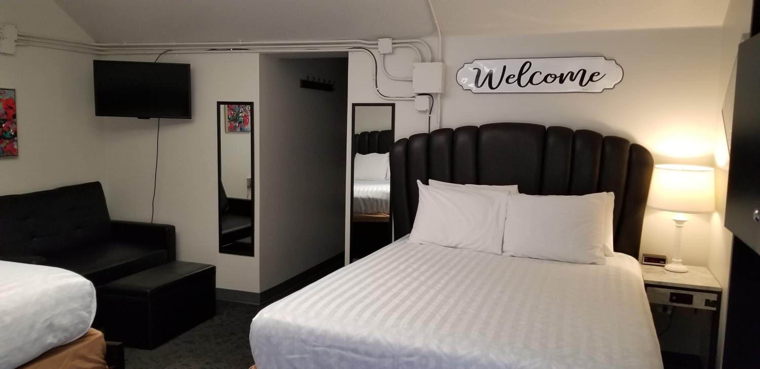 Habitación de hotel con cama, televisión, cartel de 'Welcome', sofá y mesita de noche con lámpara.