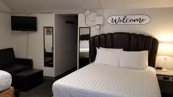 Habitación de hotel con cama, televisión, cartel de 'Welcome', sofá y mesita de noche con lámpara.