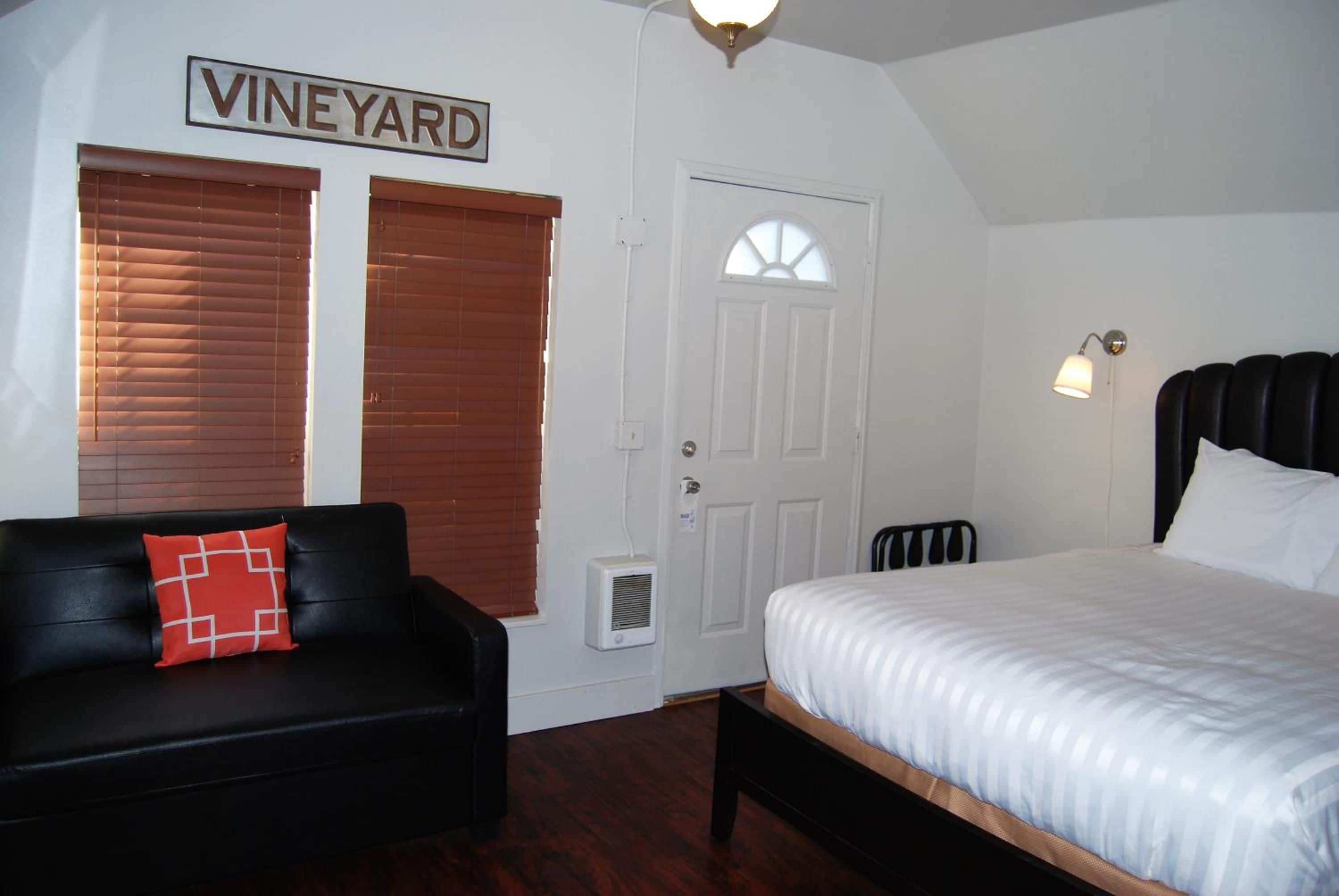 Quarto com uma cama, um sofá de couro preto com um travesseiro vermelho, uma porta branca e um letreiro na parede com a palavra 'VINEYARD'.