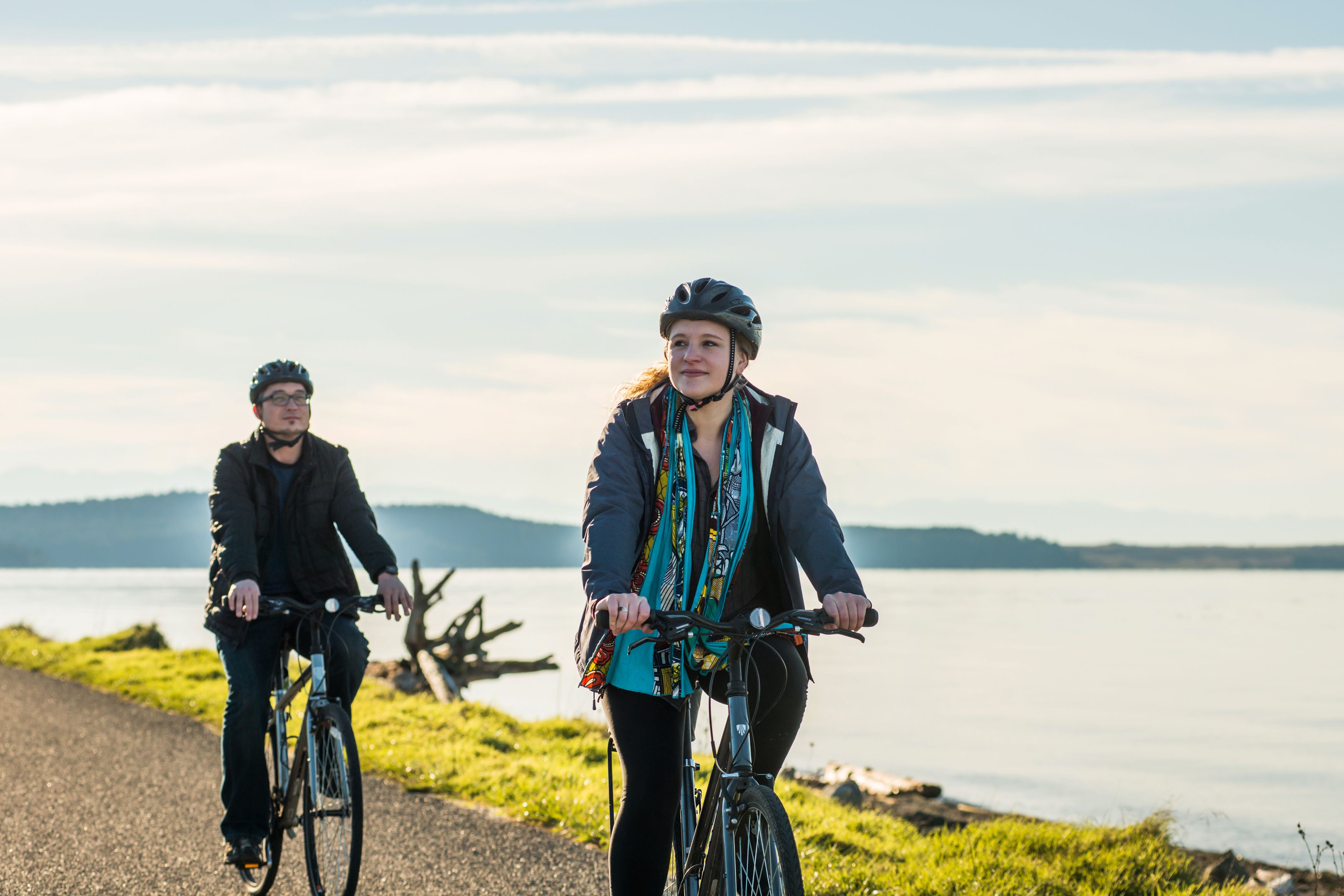 Twee personen fietsen op een pad langs een meer, met bergen op de achtergrond.