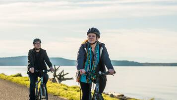Twee personen fietsen op een pad langs een meer, met bergen op de achtergrond.