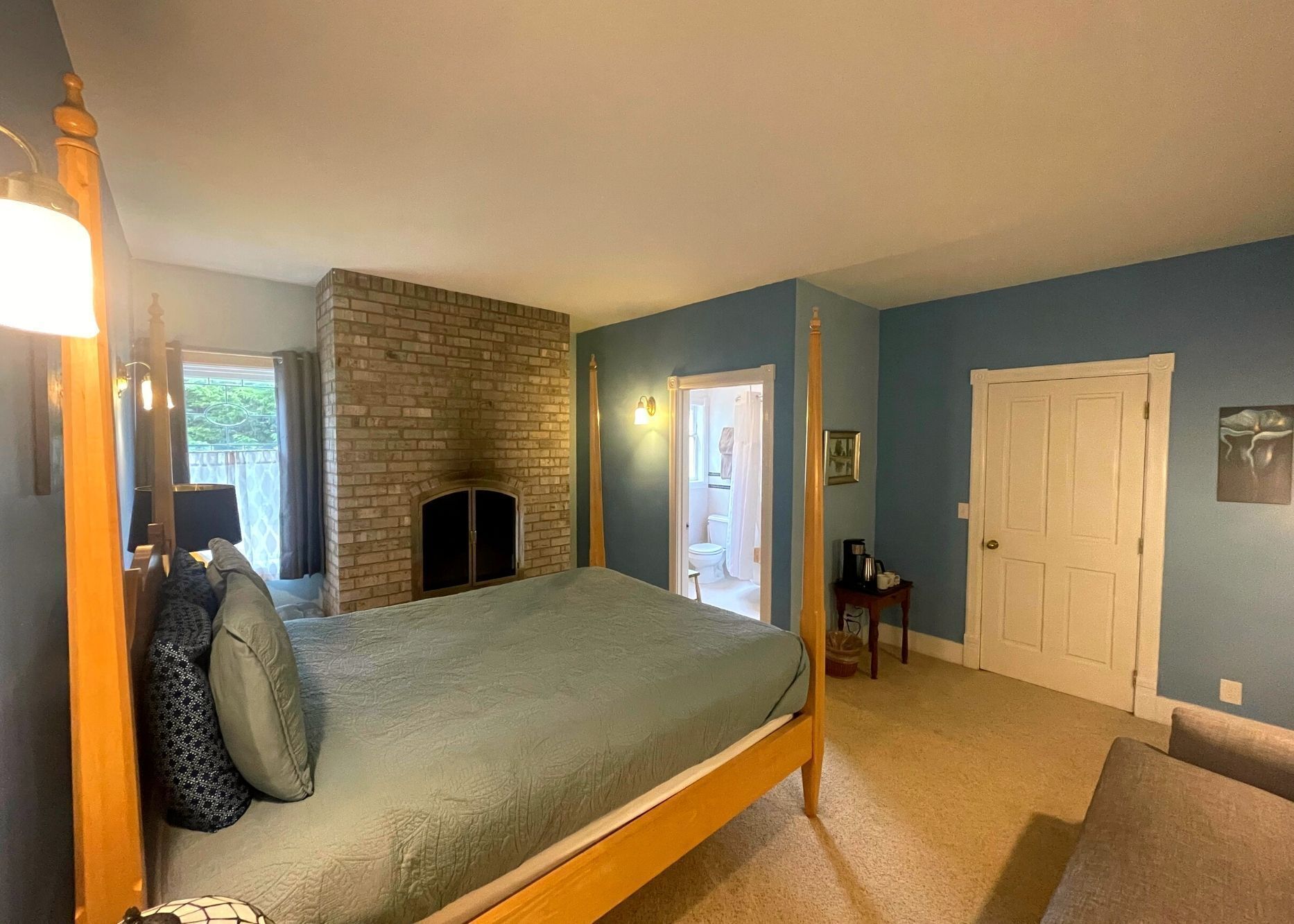 A bedroom with a bed, a brick fireplace, a door, and a small table with a coffee pot.