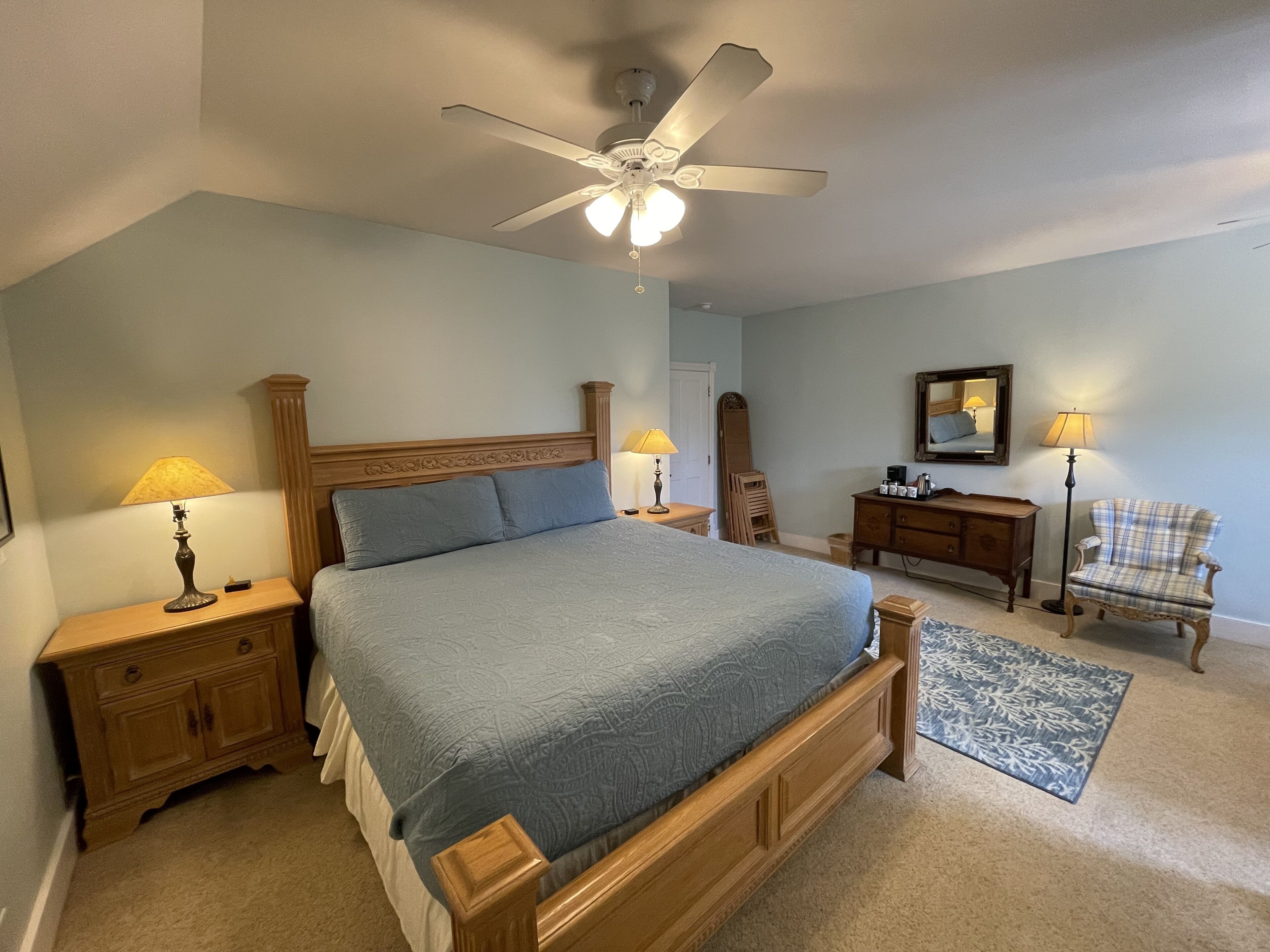 A bedroom with a large bed, wooden headboard, two bedside tables with lamps, a ceiling fan, a dresser, a chair, and a mirror on the wall.