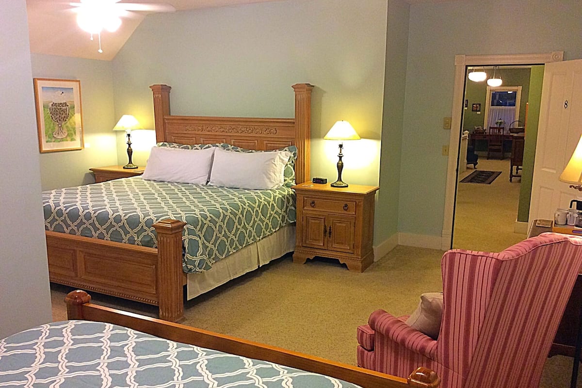 A bedroom with a bed, two bedside tables with lamps, a chair, and a door leading to another room.