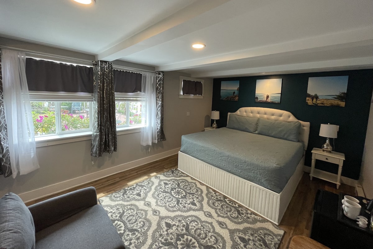 A bedroom with a large bed, a window with curtains, a bedside table with a lamp, and a patterned rug.
