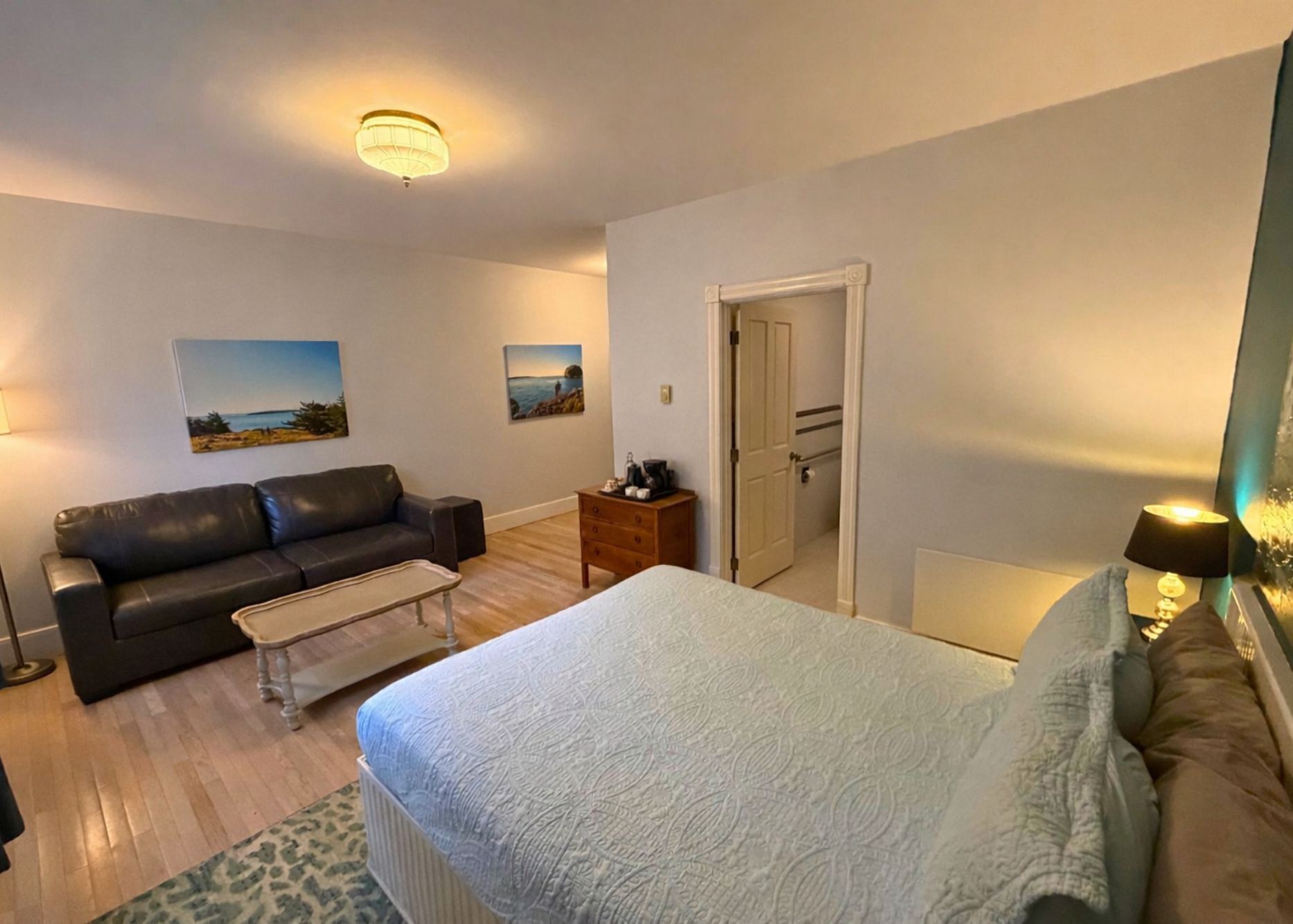Quarto com cama, sofá, mesa de centro e cômoda. Há duas pinturas na parede e uma porta que dá para outro cômodo.