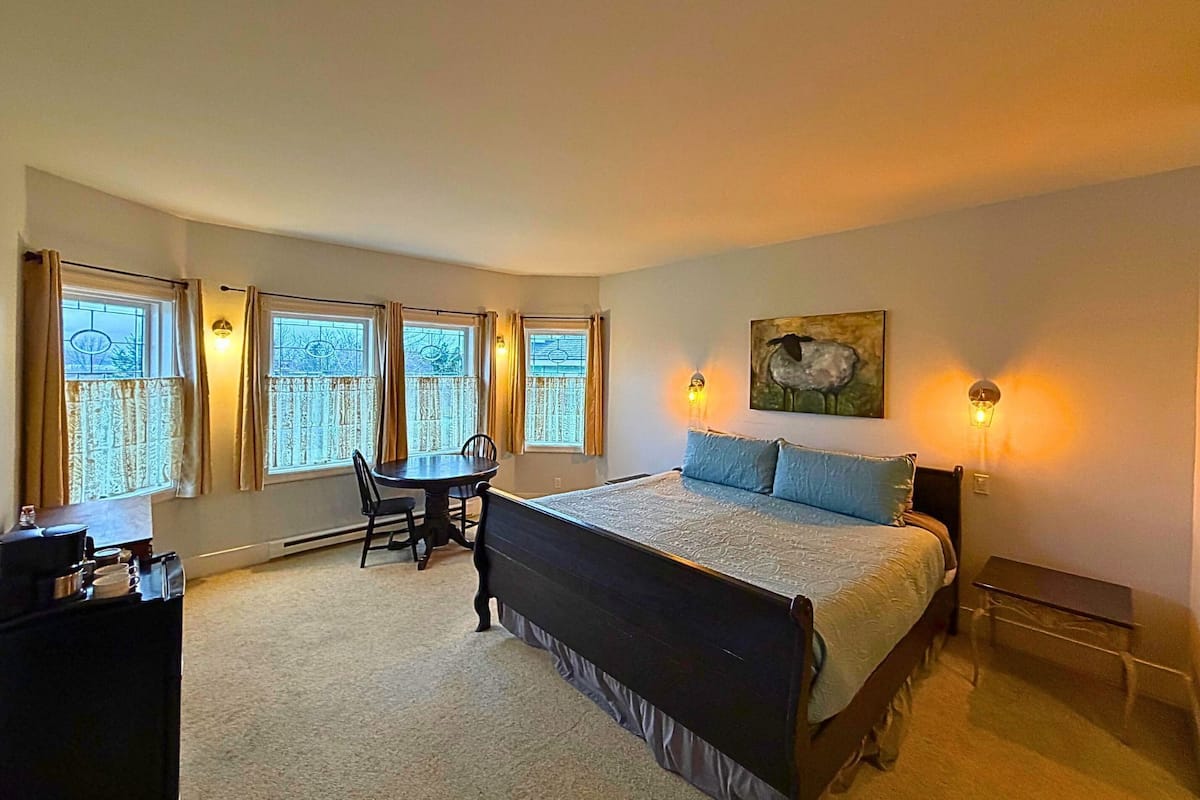 A bedroom with a bed, a desk, and a chair. There are windows with curtains, a painting on the wall, and wall sconces.