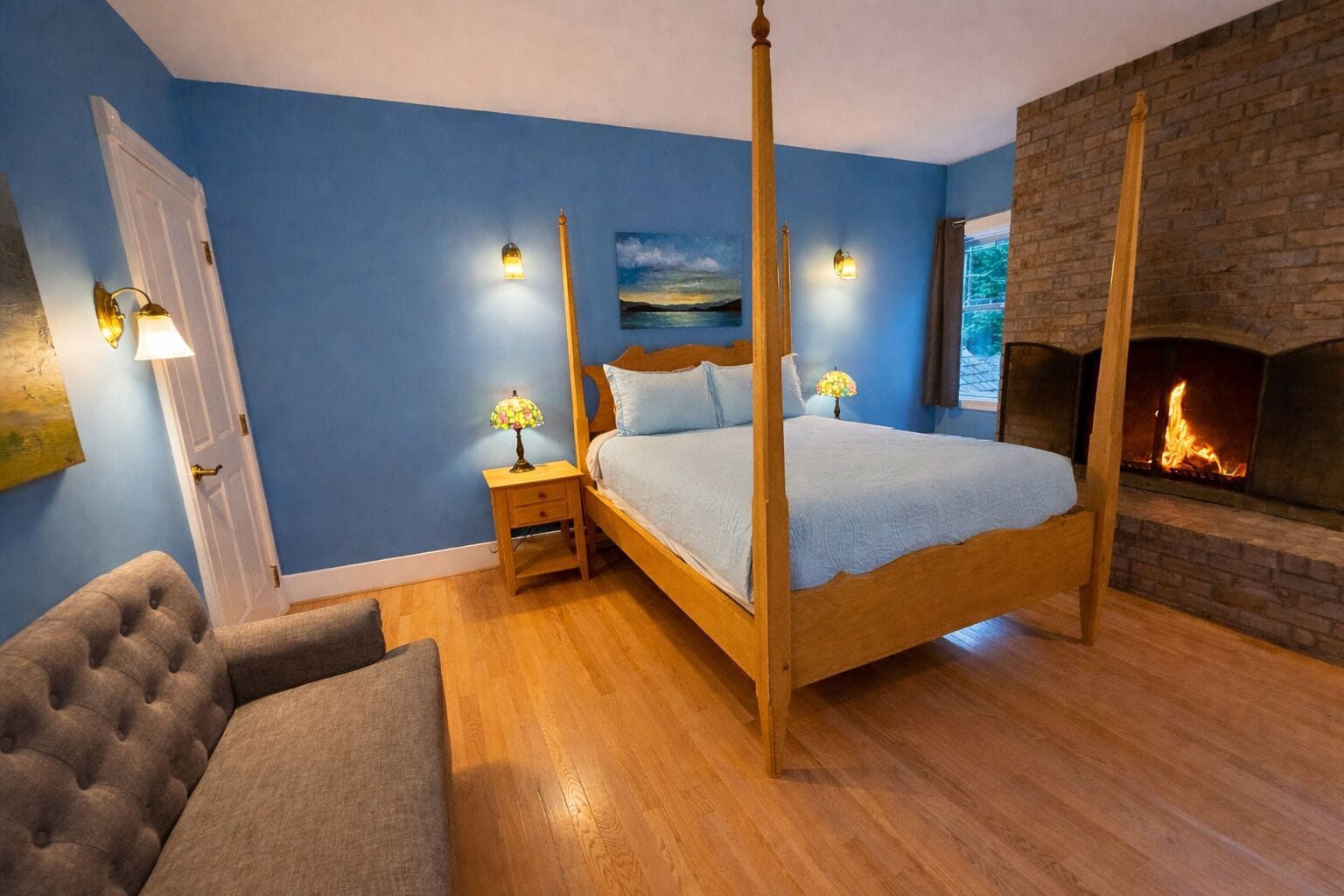 A four-poster bed with a headboard, two bedside tables with lamps, a fireplace, and a tufted armchair.