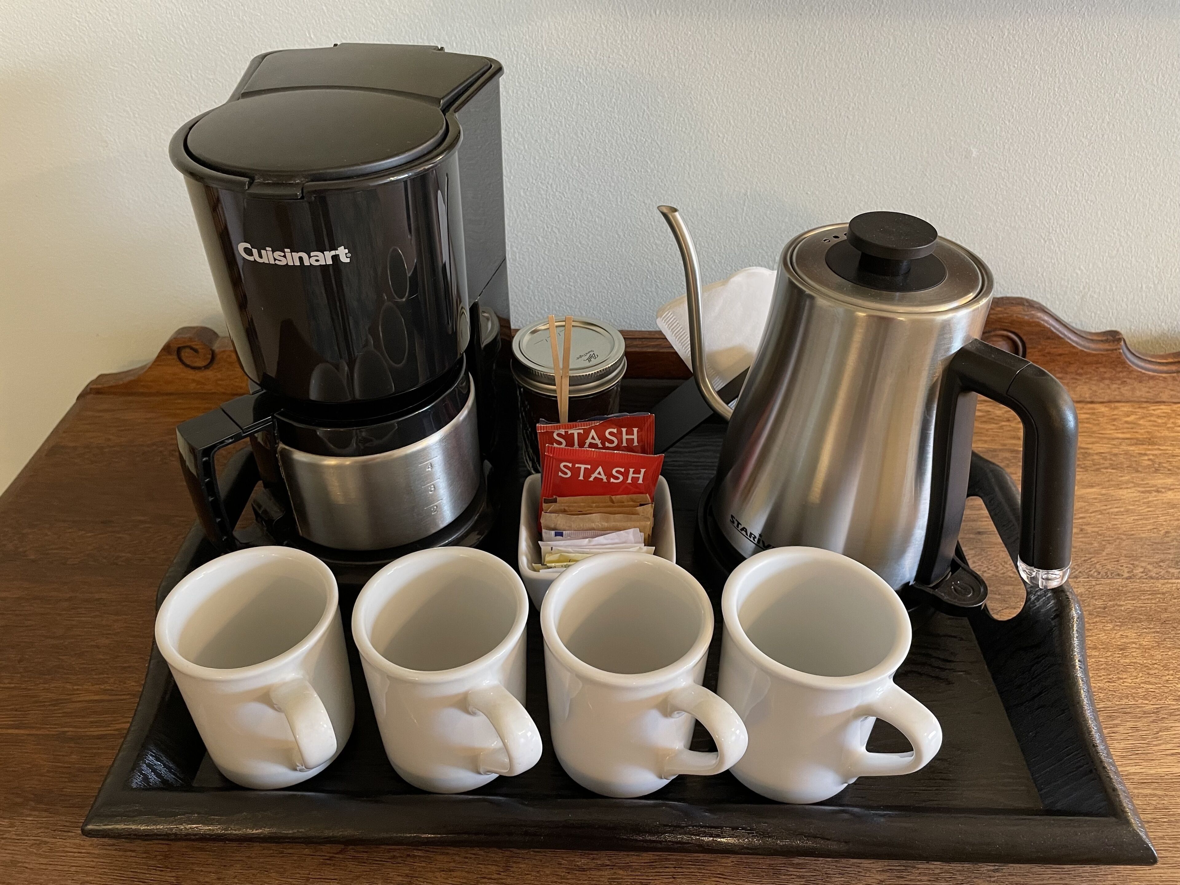 A coffee maker, a stainless steel pot, a jar labeled "STASH STASH," and four white mugs on a tray.