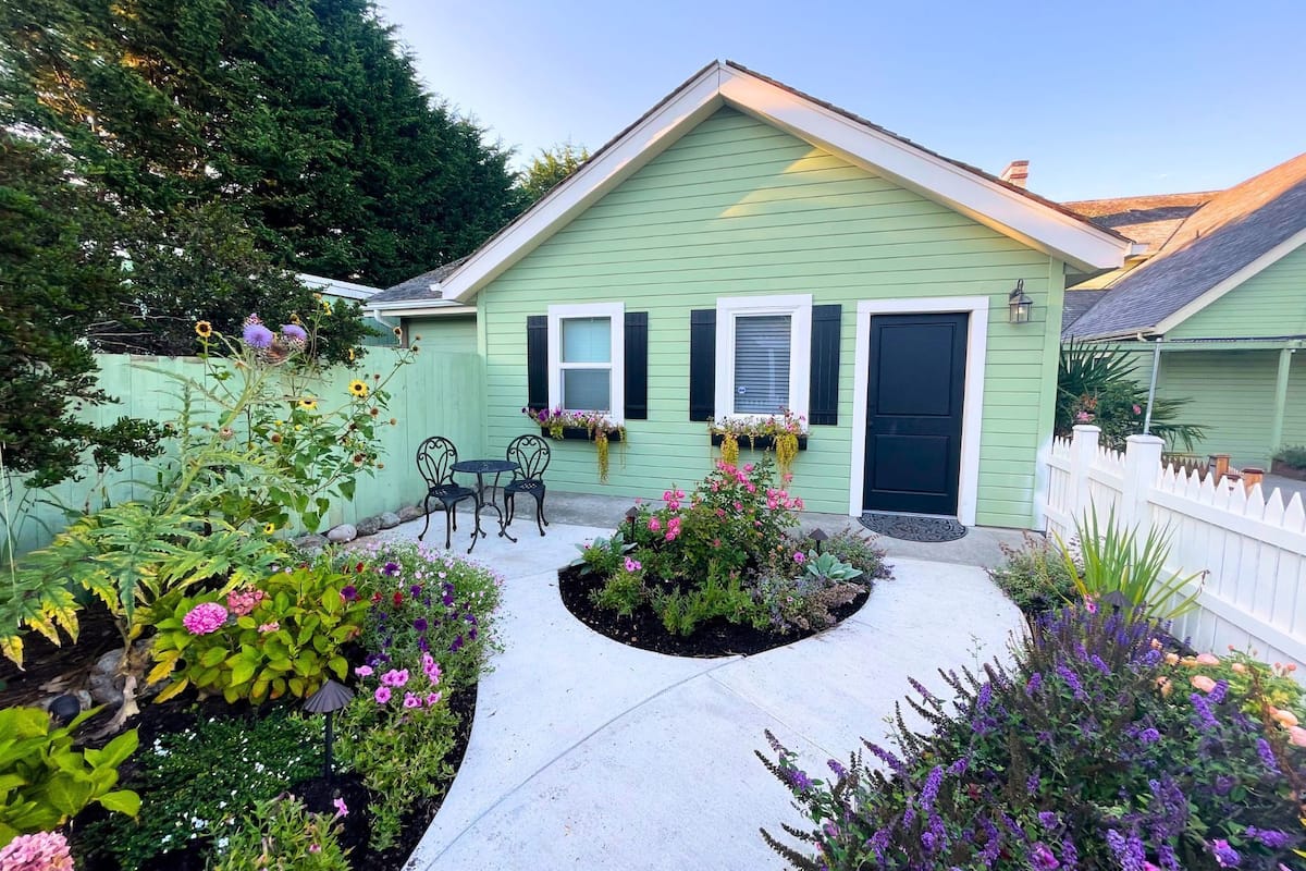 A green house with a front porch, two windows, and a door. There is a small garden with various plants and flowers.