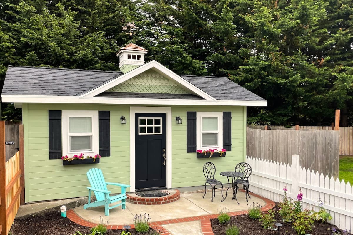 A small house with a green exterior, a black door, and two windows with shutters. There is a blue Adirondack chair and a small table with chairs outside.
