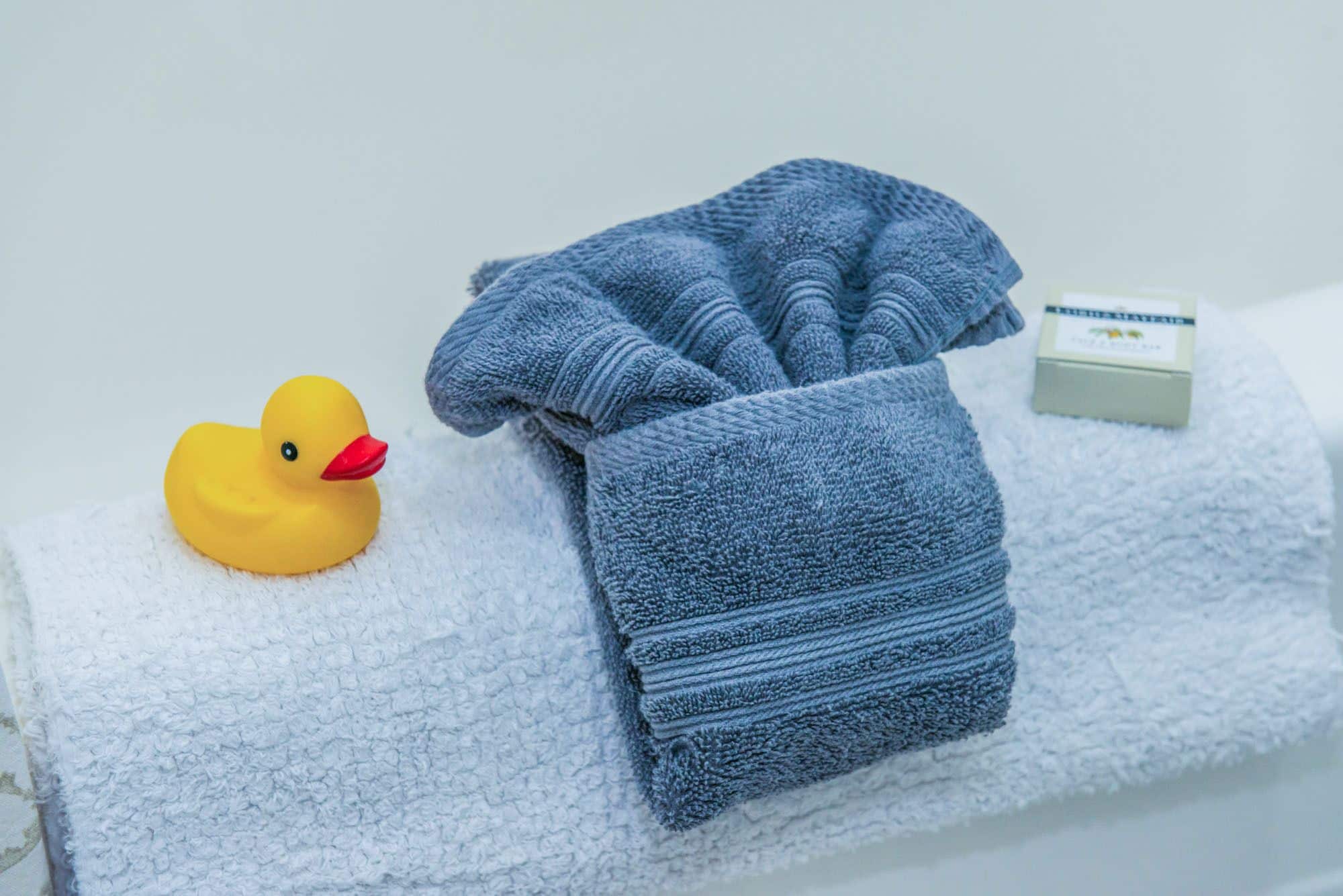 A white towel, a folded blue towel, a yellow rubber duck, and a small white box with a green label.