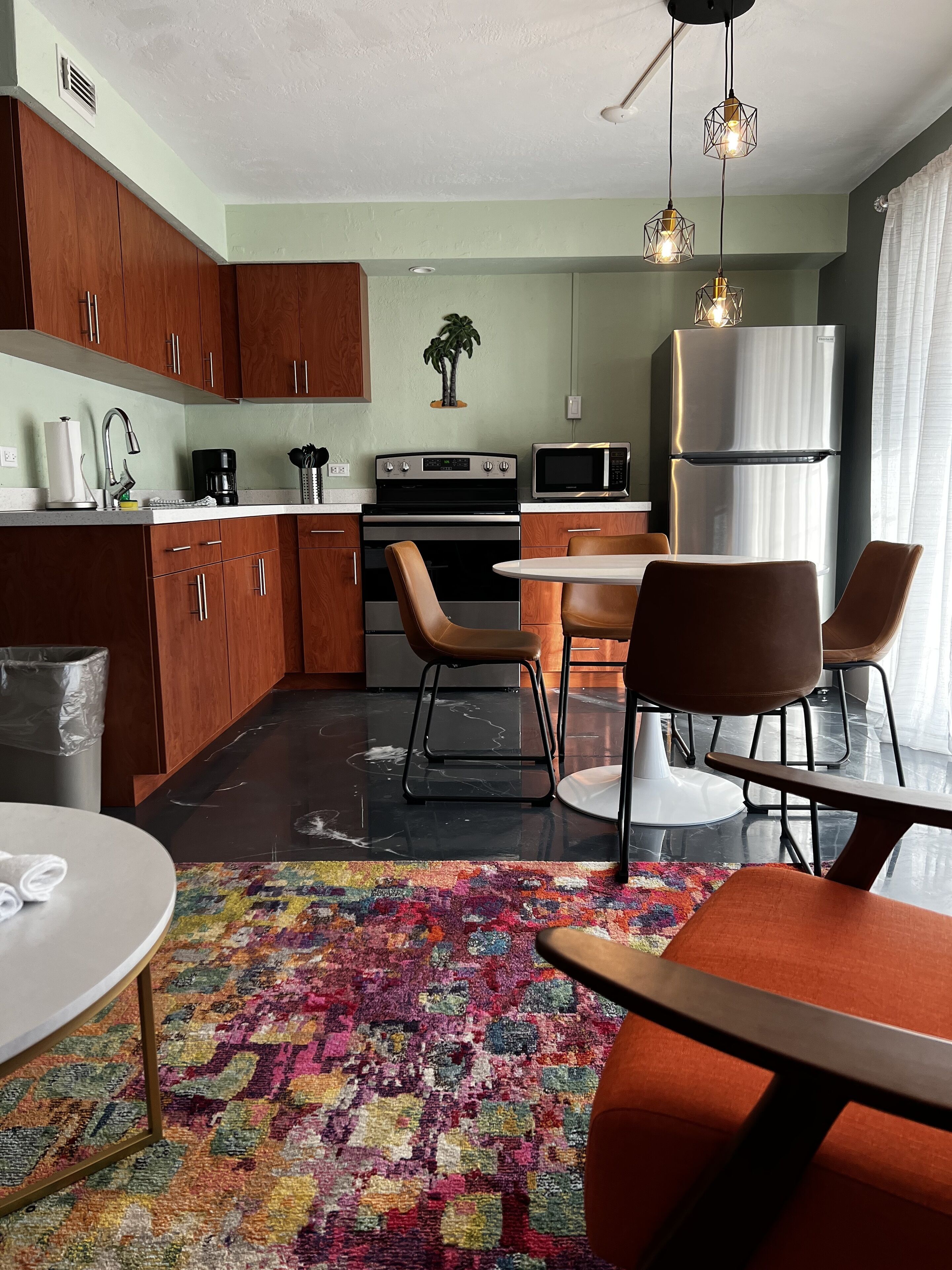 A modern kitchen with wooden cabinets, a stainless steel refrigerator, a microwave, and a stove. There is a dining area with a table and chairs, and a colorful rug on the floor.