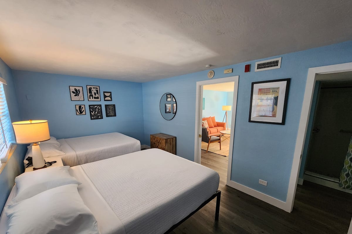 Une chambre avec deux lits, une table de chevet avec une lampe et une porte donnant sur une autre pièce.