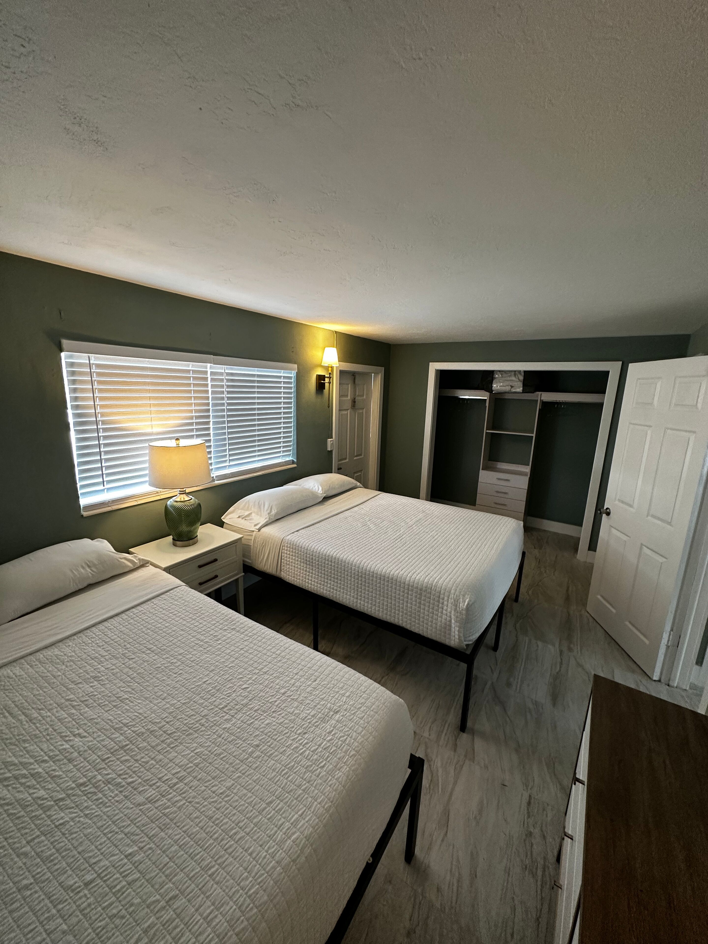 A room with two beds, a nightstand, a lamp, a window with blinds, a closet, and a door.