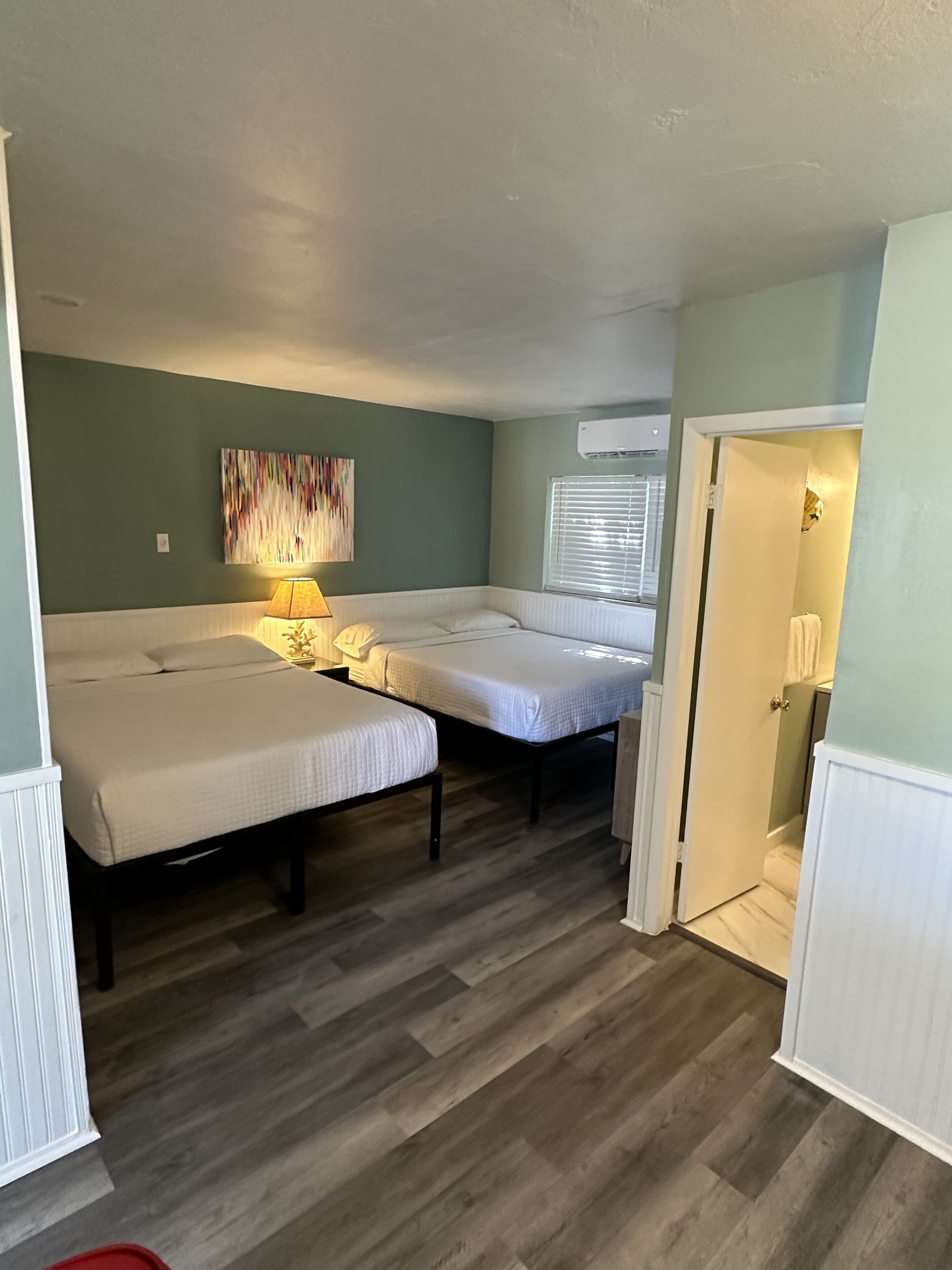 Habitación compacta con dos camas individuales, una mesita con lámpara, un cuadro abstracto de colores en la pared y una ventana con persianas.