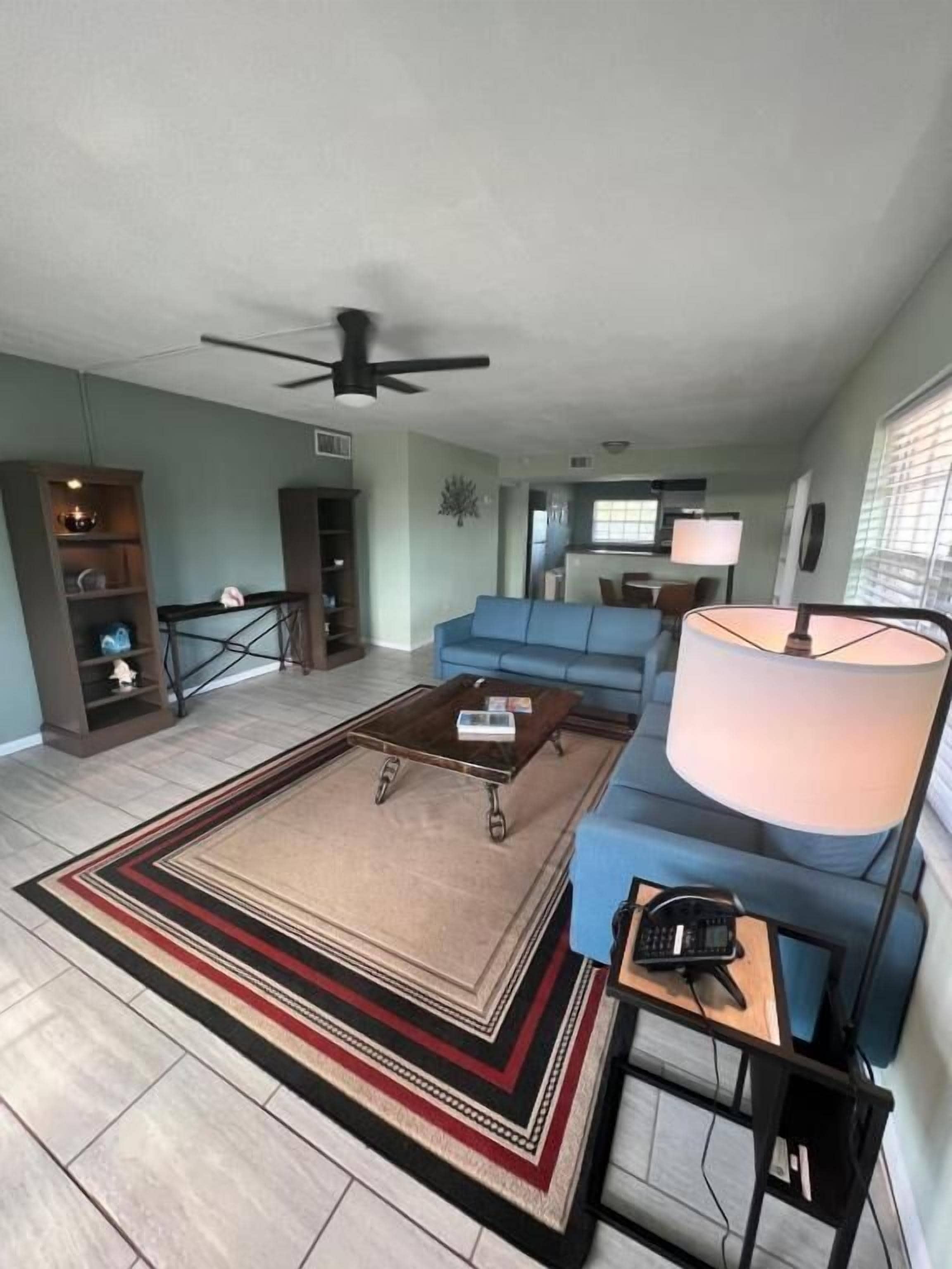 A living room with a sofa, coffee table, and lamp. There is a ceiling fan and a television in the background.