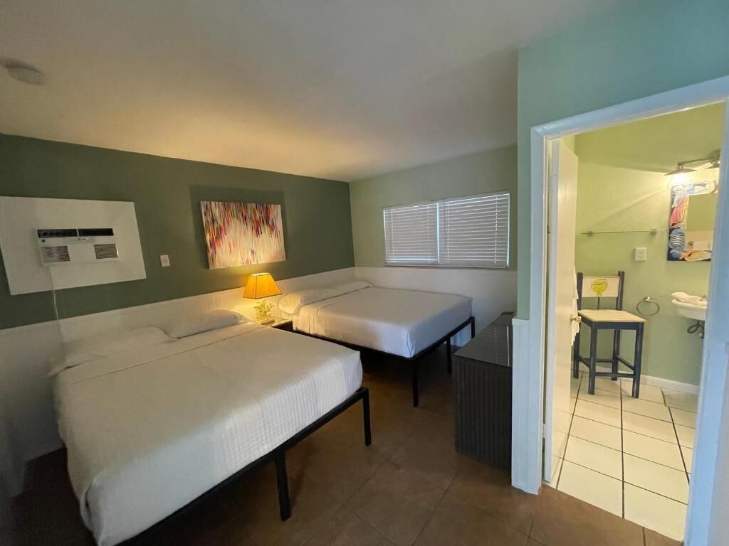 Une chambre avec deux lits, un climatiseur fixé au mur, une fenêtre avec des stores et une salle de bain visible à travers une porte ouverte.