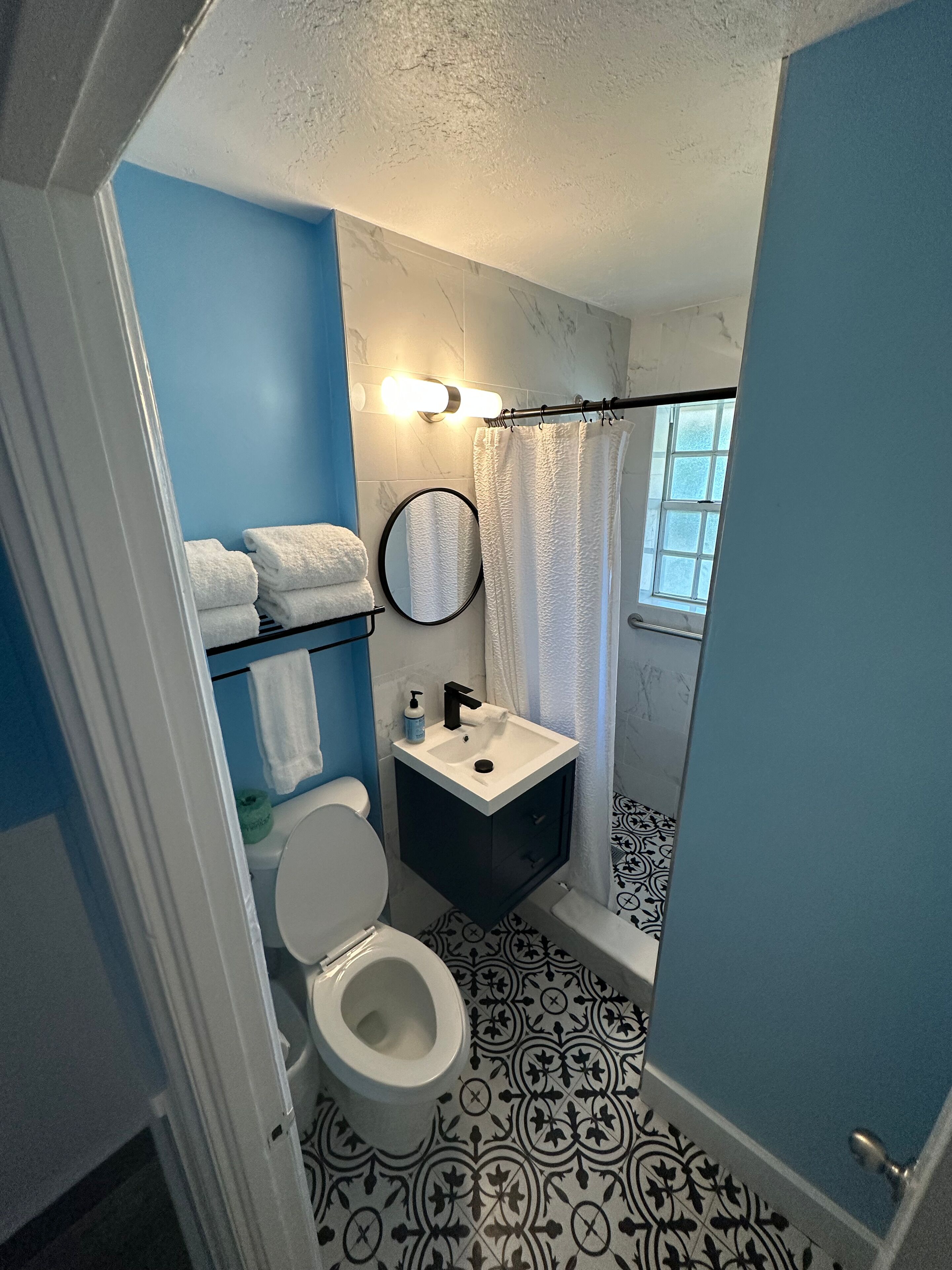 A bathroom with a toilet, sink, and shower curtain. There are towels on a rack and a window providing natural light.
