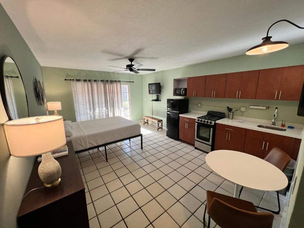 A compact room with a bed, a desk with a lamp, a small kitchen area with a stove and microwave, and a dining table.