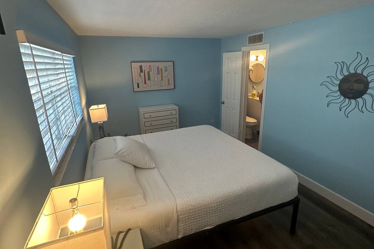 Une chambre à coucher comprenant un lit, une table de chevet, une lampe et une œuvre d’art accrochée au mur.