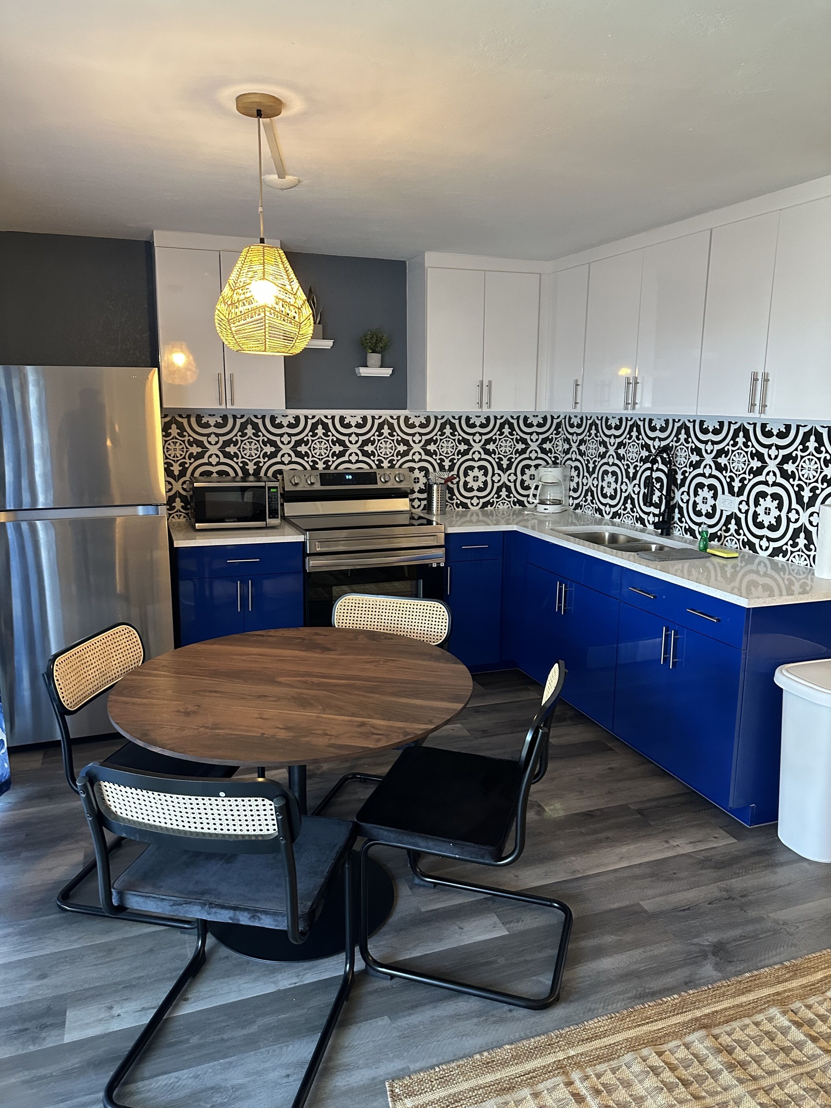 Una cucina moderna con mobili blu, un frigorifero in acciaio inossidabile, un forno a microonde e un forno. È presente un tavolo rotondo in legno con due sedie e una lampada a sospensione sopra.