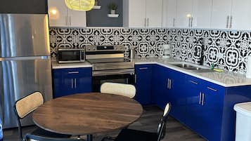 Una cucina moderna con mobili blu, un frigorifero in acciaio inossidabile, un forno a microonde e un forno. È presente un tavolo rotondo in legno con due sedie e una lampada a sospensione sopra.