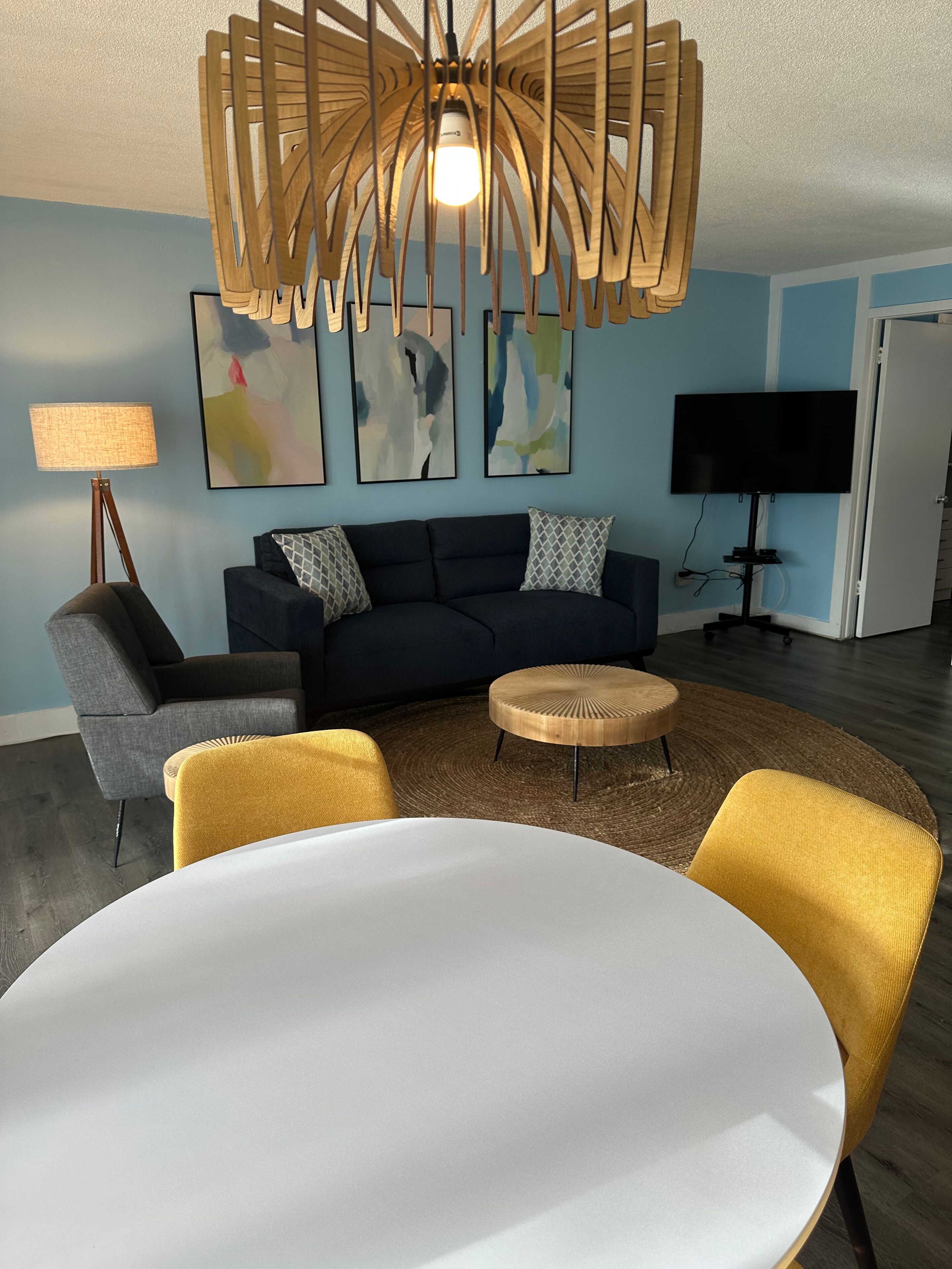 A modern living room with a round white table, a dark blue sofa, and a yellow armchair. There is a unique wooden ceiling light fixture, a flat-screen TV, and abstract wall art.