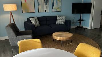 A modern living room with a round white table, a dark blue sofa, and a yellow armchair. There is a unique wooden ceiling light fixture, a flat-screen TV, and abstract wall art.