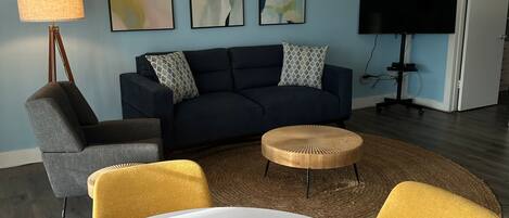 Una sala de estar moderna con una mesa redonda blanca, un sofá azul oscuro y un sillón amarillo. Se observa una lámpara de techo de madera original, un televisor de pantalla plana y arte abstracto en la pared.
