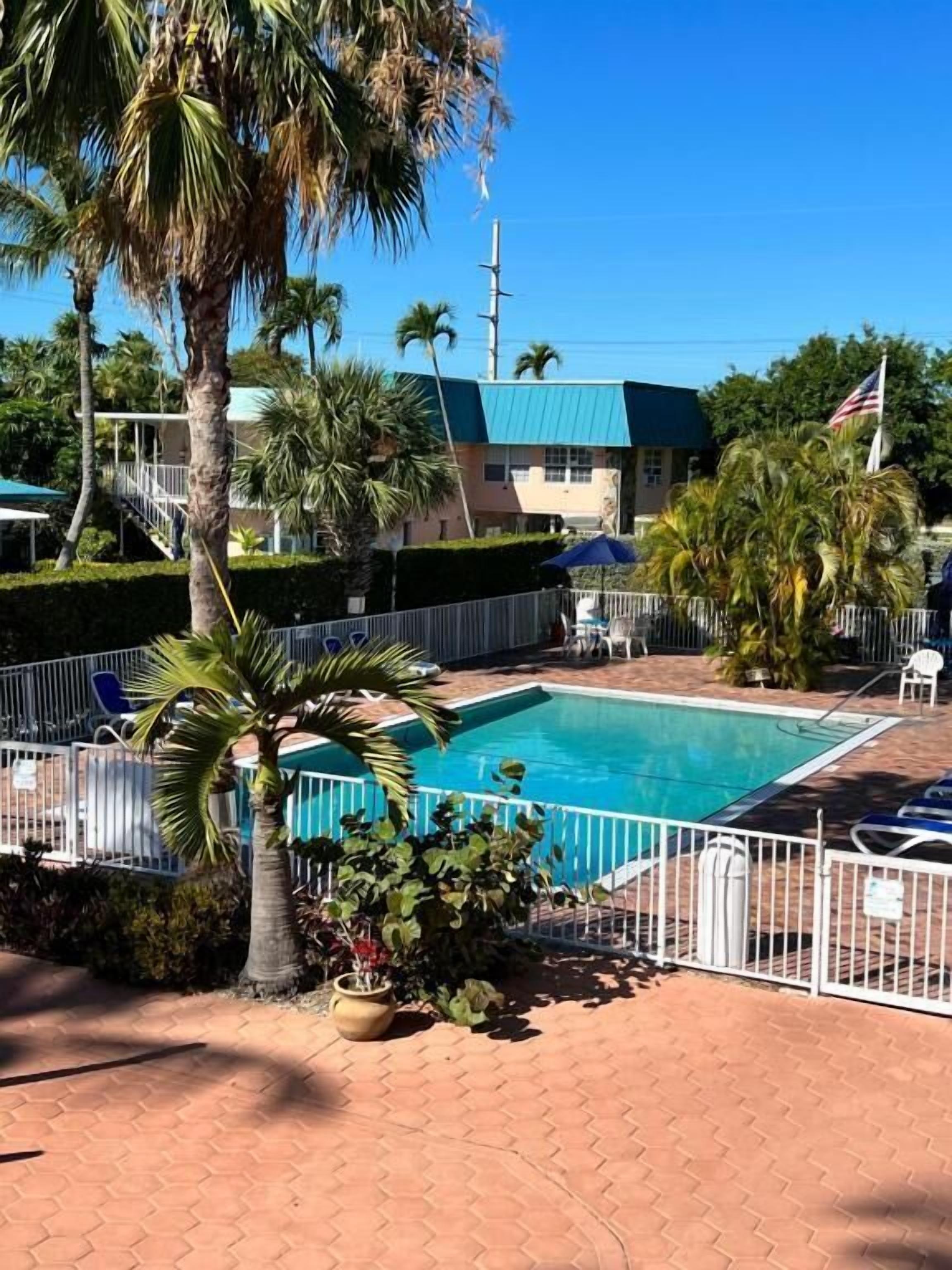 A swimming pool with a fenced area, palm trees, and a building with a green roof in the background.