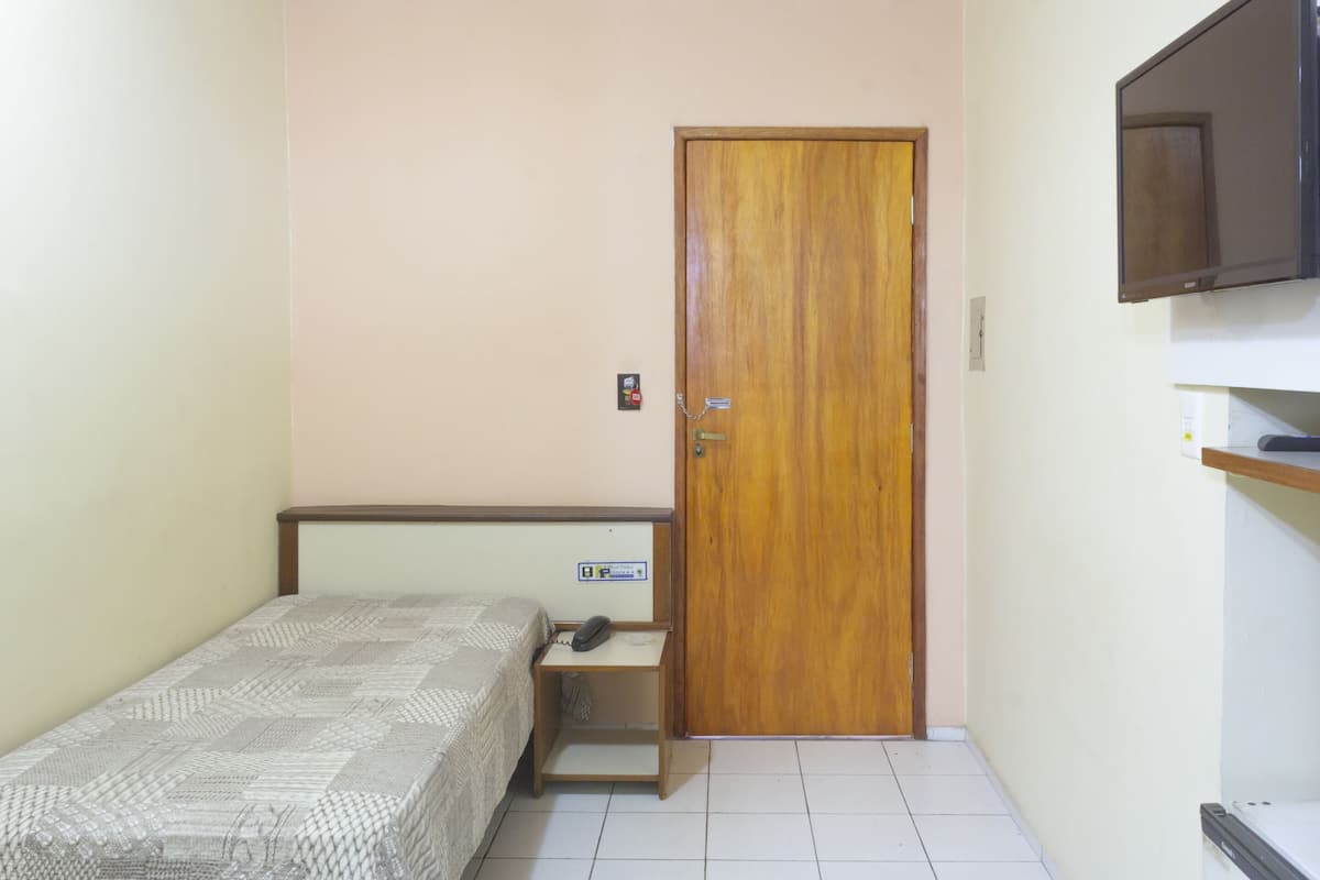 A small, neatly arranged bedroom with a single bed, a bedside table with a phone, a wooden door, and a mounted TV.